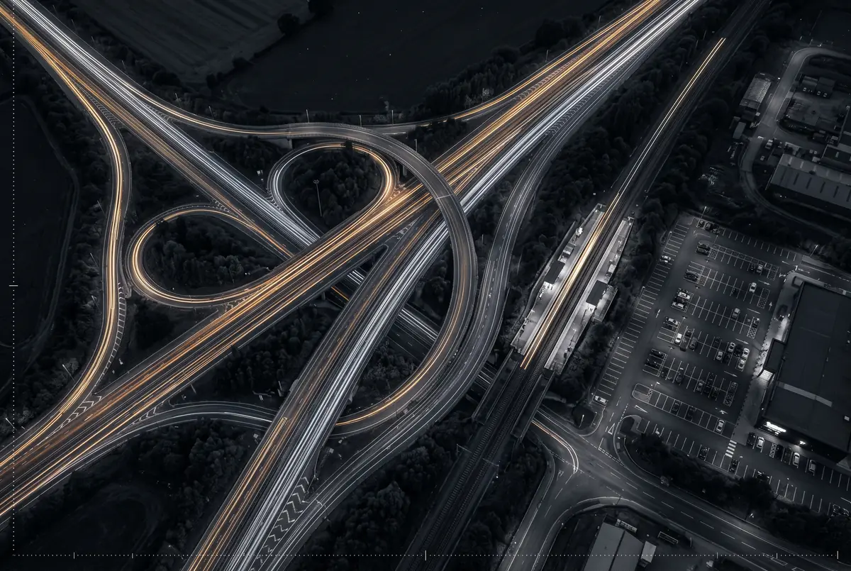 KI-Illustration: ein Autobahnkreuz mit Bahnlinie und Lichtspuren bei Nacht (kein realer Ort)